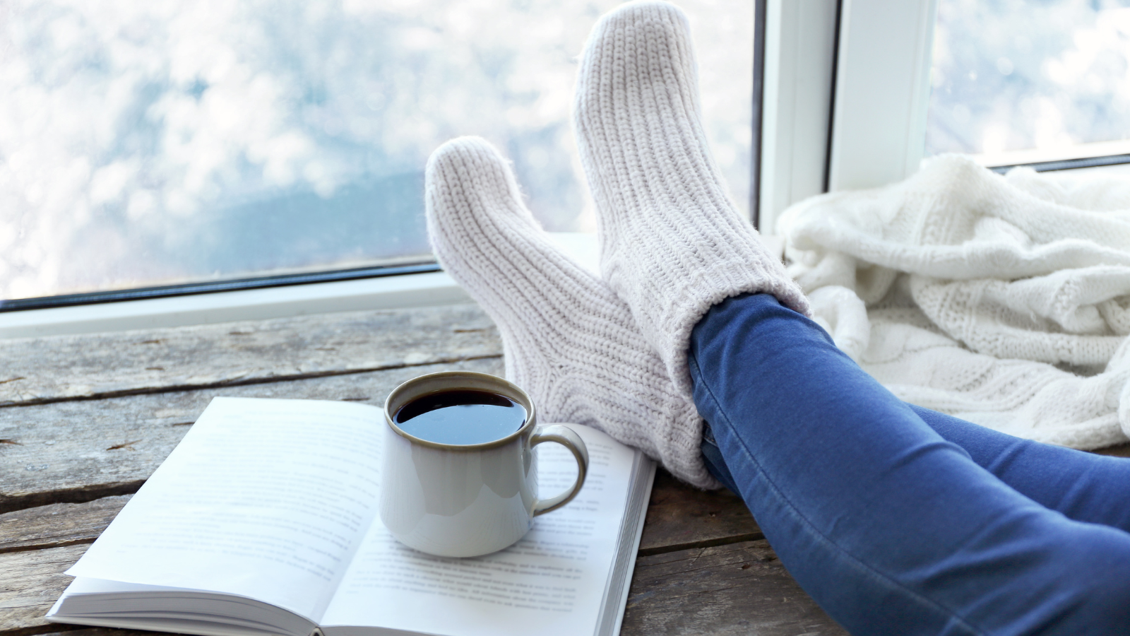 Womans legs crossed on table with warm socks on and a book with coffee