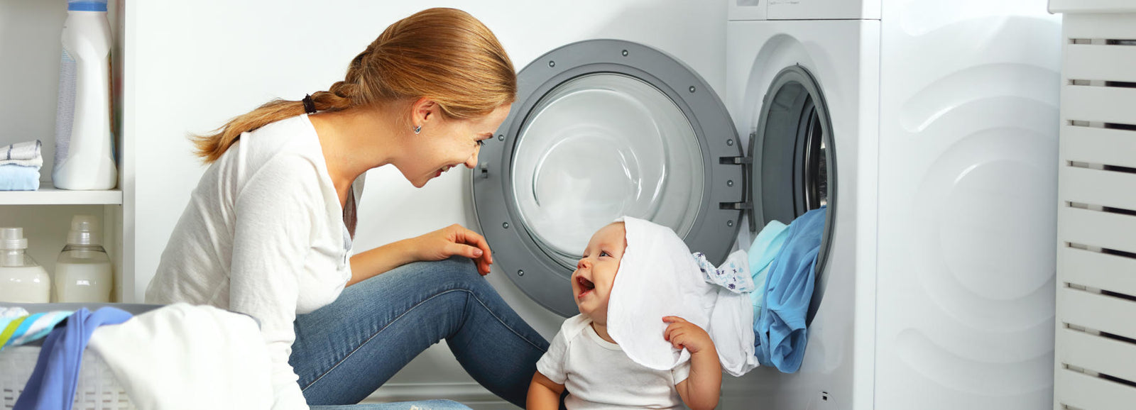 A woman sits on the floor smiling at a baby in front of an open washing machine filled with laundry.